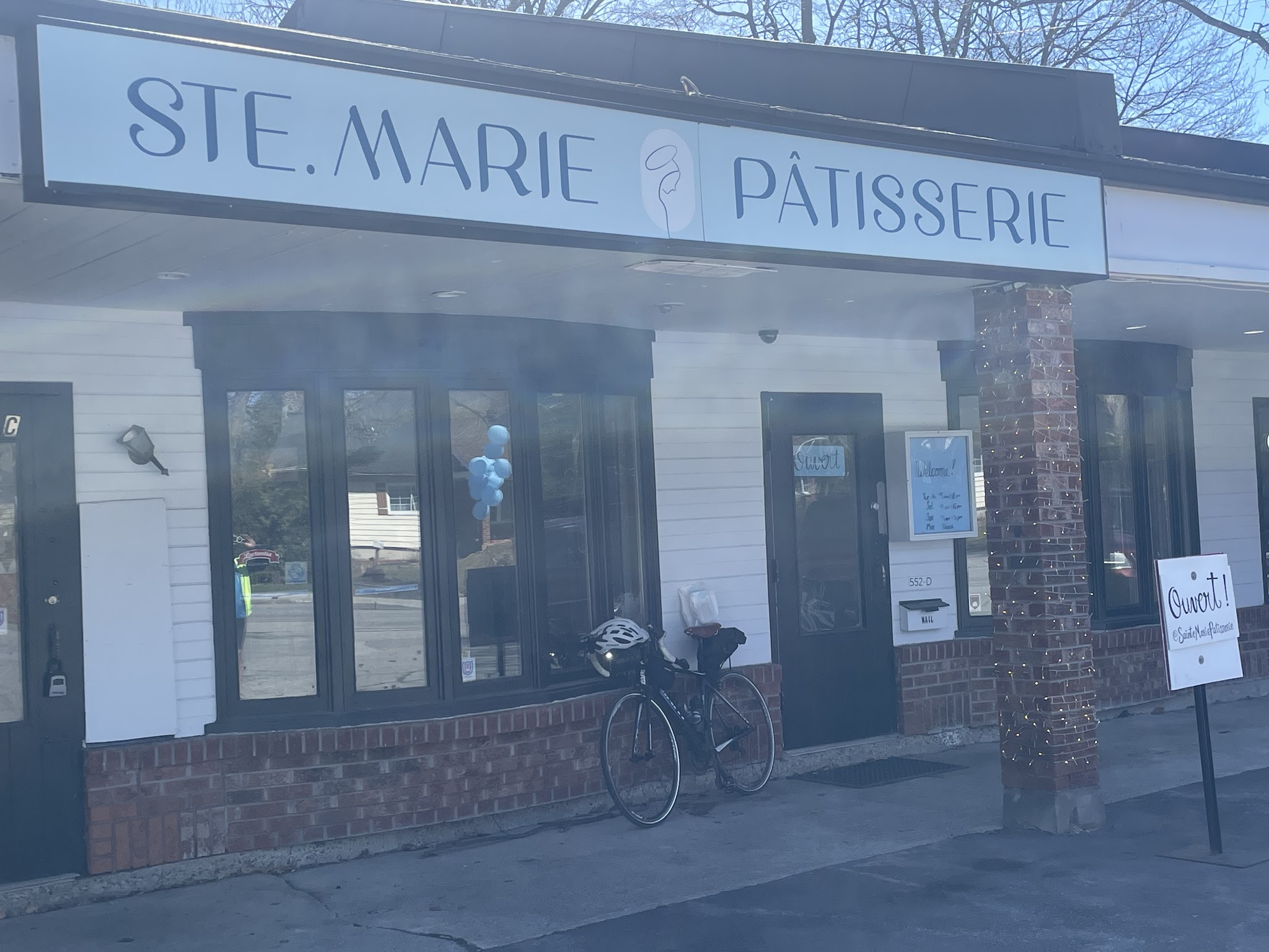 Daytime exterior of Sainte Marie Pâtisserie with signage, bicycle parked outside, blue balloons, and welcome sign