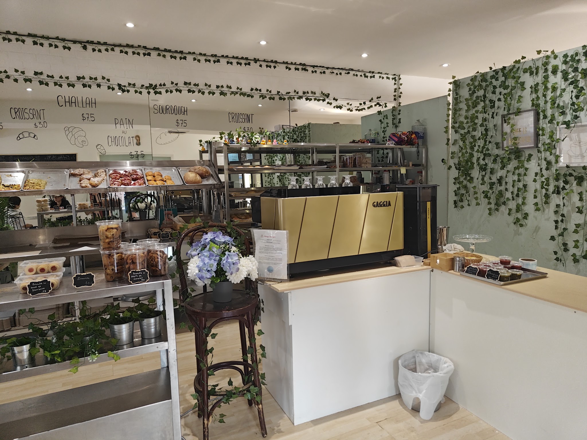 Interior of Sainte Marie Pâtisserie showing the counter area with a gold Gaggia espresso machine, ivy-draped walls, chalkboard menu listing croissants, challah, sourdough, and pain au chocolat